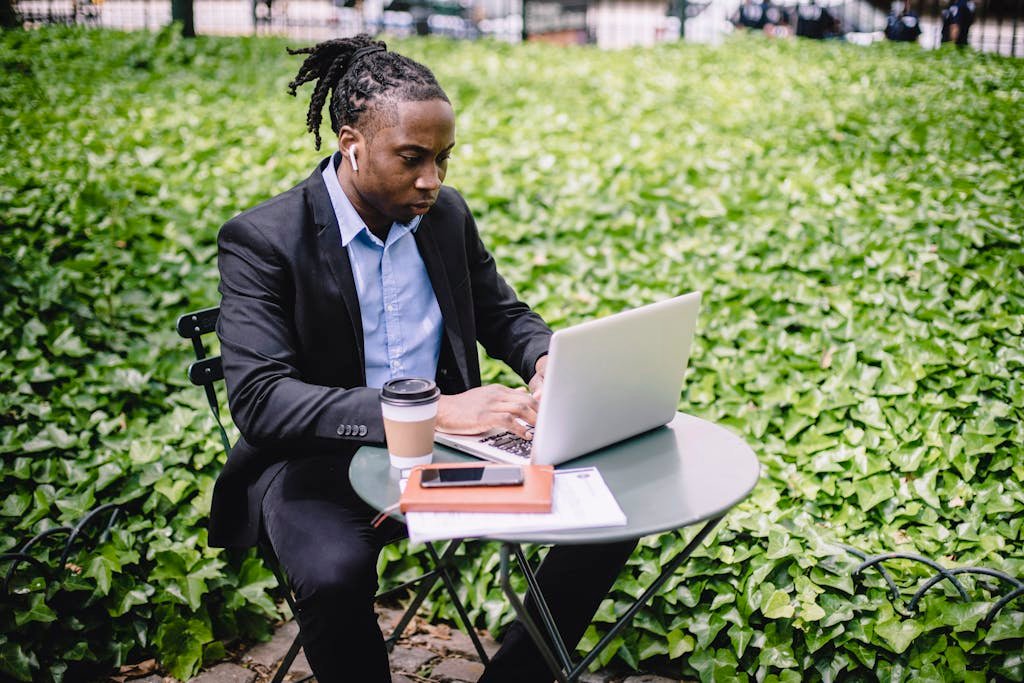 Concentrated African American businesswoman in earbuds wearing formal black suit sitting at table in city garden and typing on netbook