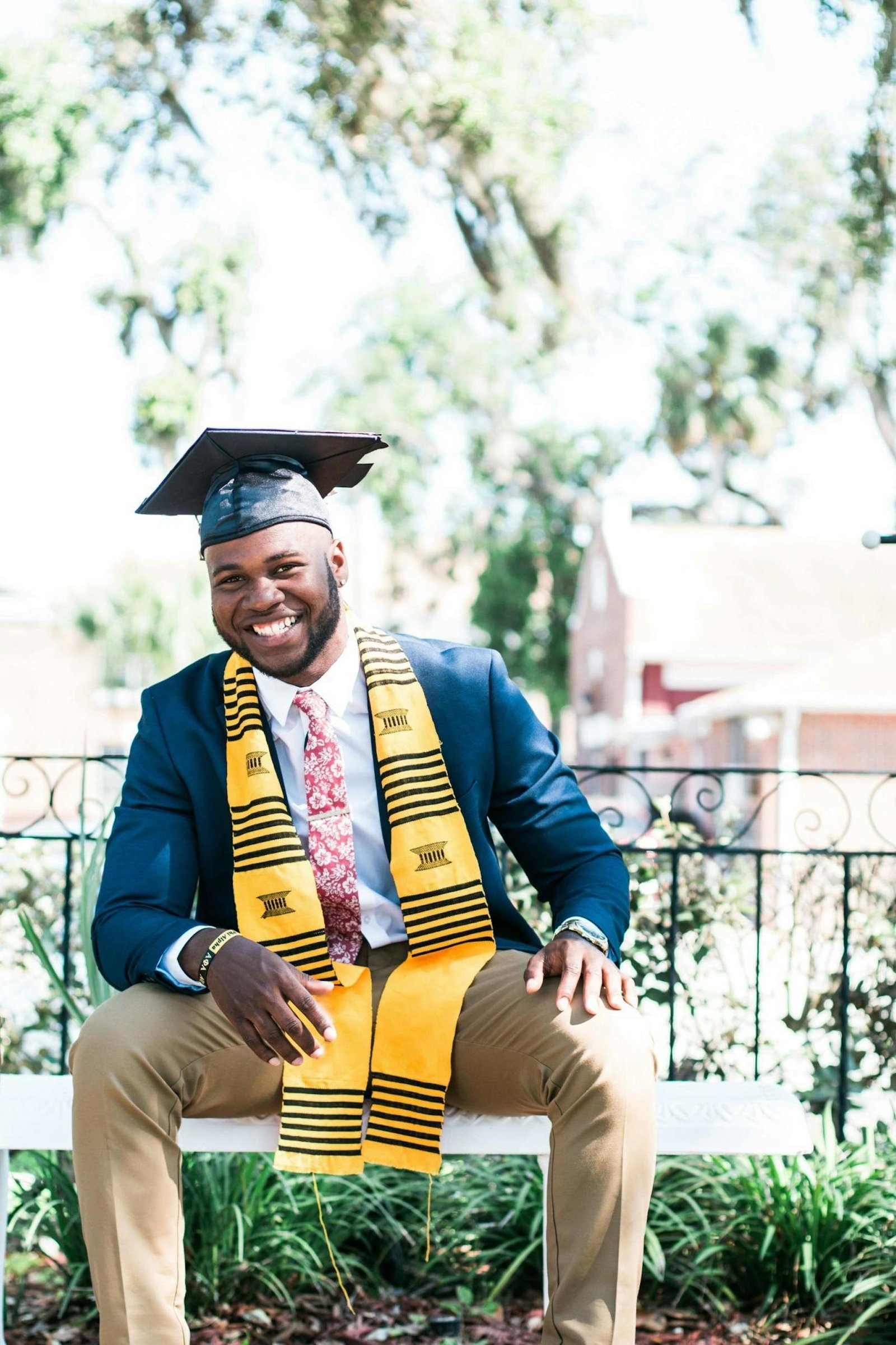 Smiling graduate sitting outside wearing a cap and gown with stole.